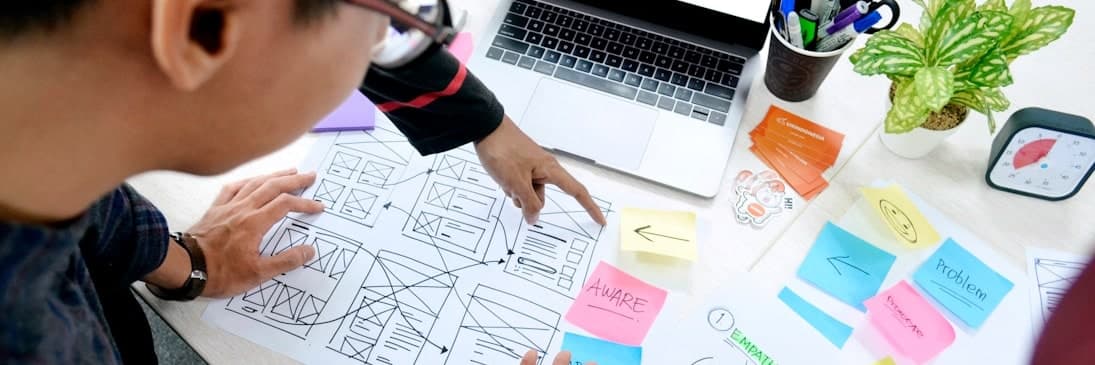 Dos personas revisan wireframes impresos sobre un escritorio, junto a una notebook y notas adhesivas de colores